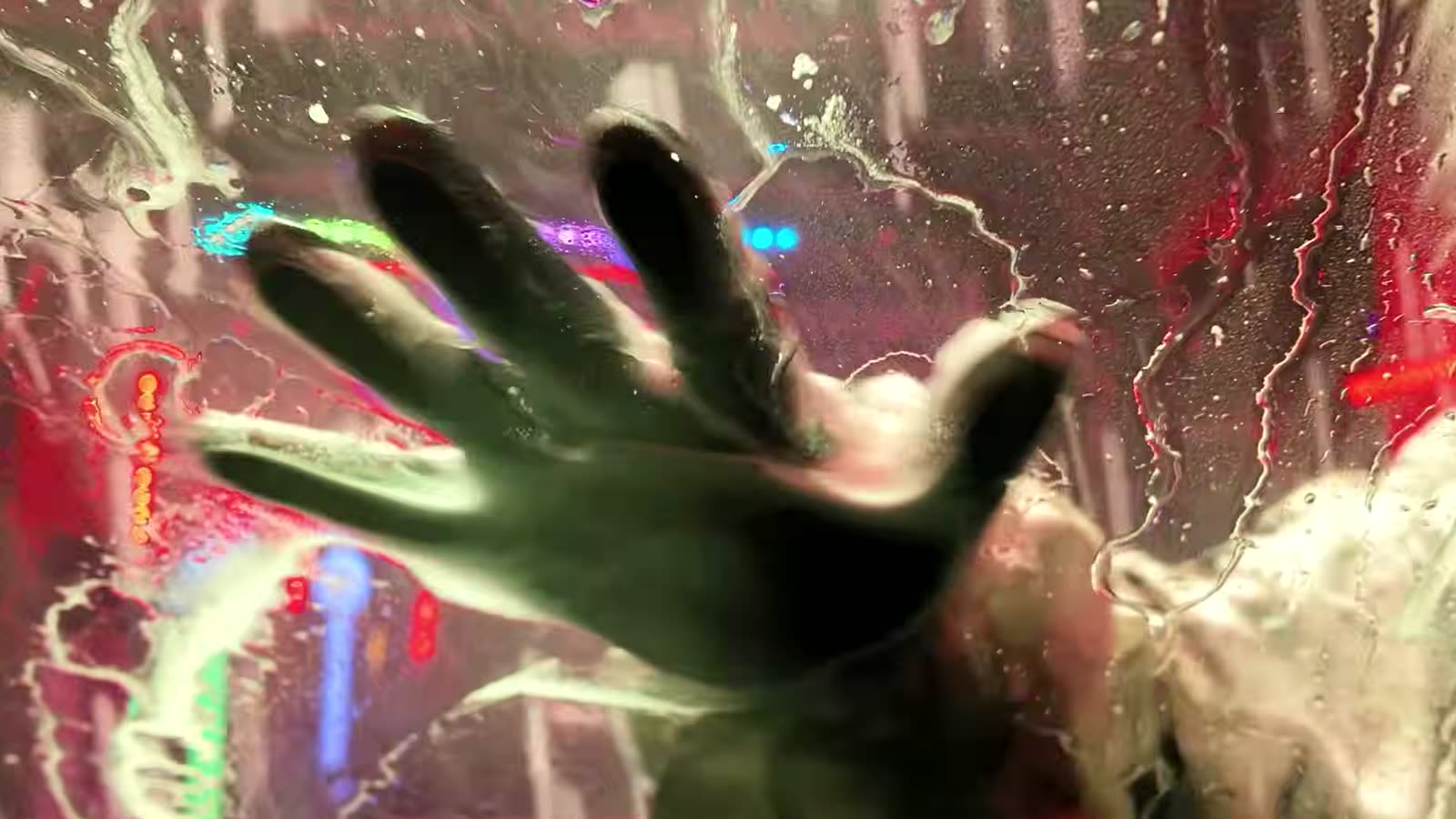 A gloved hand pressed flat against a soapy windscreen, lit by coloured tunnel lights.