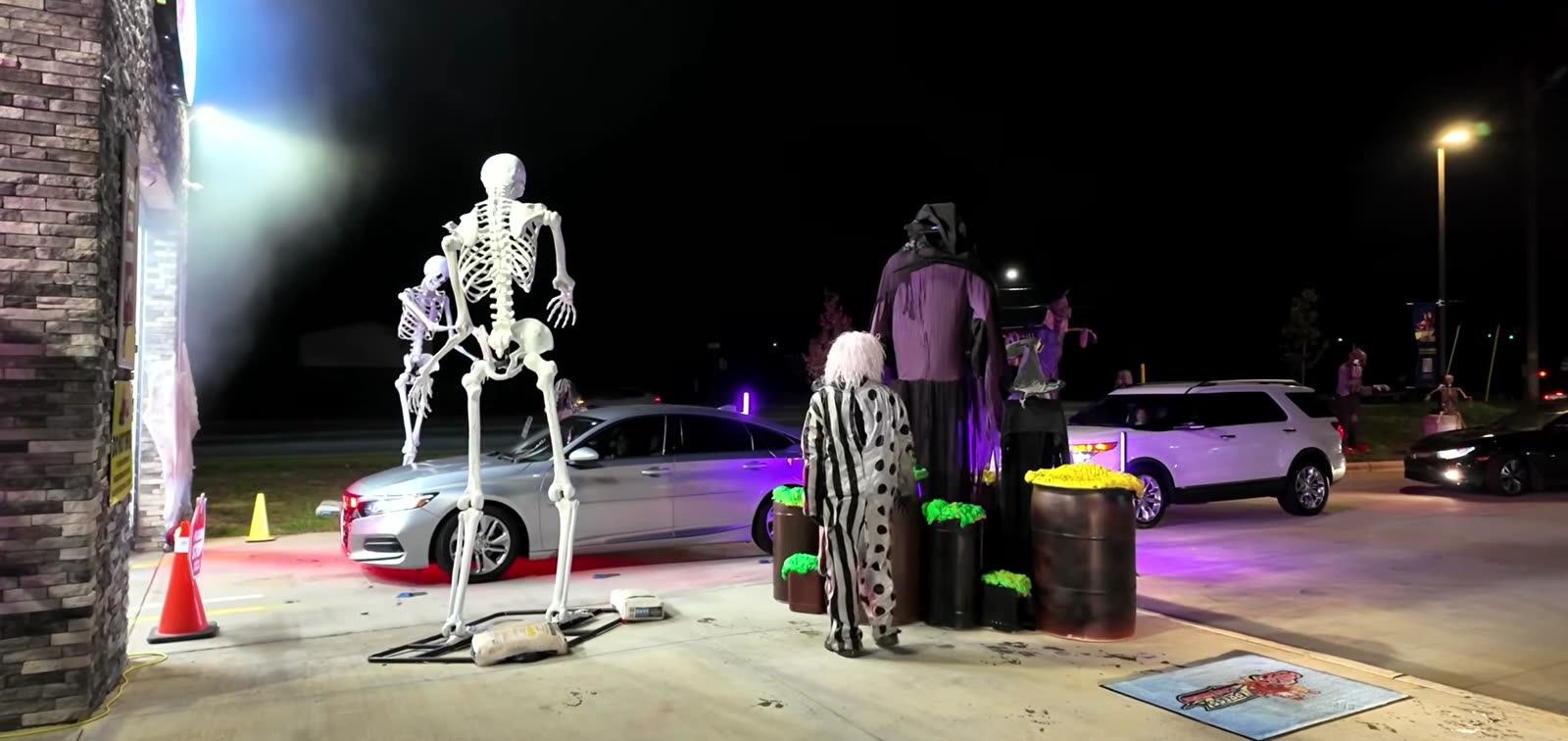 A carwash lot at night during the event, lit purple, with tall Halloween props, costumed staff and guest vehicles queuing.
